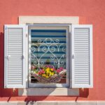Close-up of a window on a coral-colored building with white open shutters and a flower basket with yellow and pink daisies.
