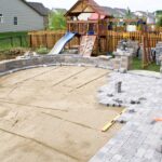 A patio area of a fenced-in yard remains partially completed as sand and stone bricks cover portions of the area.