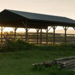 An old pole barn is in front of a fence and a pile of logs with the sun and mountains in the background.