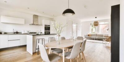 A dining table with six gray chairs stands in front of a kitchen with white cabinets and black countertops.