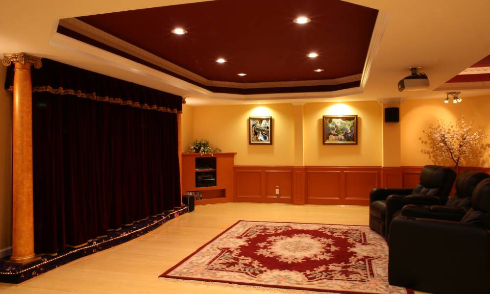 Three leather recliners sit on a red oriental rug in a home theater with recessed lighting and a velvet curtain.