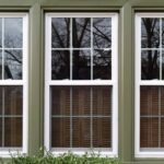Tips To Enhance the Window Protection of Your Home Three large vertical sliding windows with white trim and a green frame reflect the image of tree branches.