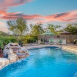 A circular backyard pool with a stone water feature surrounded by landscaping and two chairs and umbrellas.