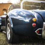 A rear view of a blue vintage convertible with white racing stripes, chrome accents, and round taillights parked on grass.