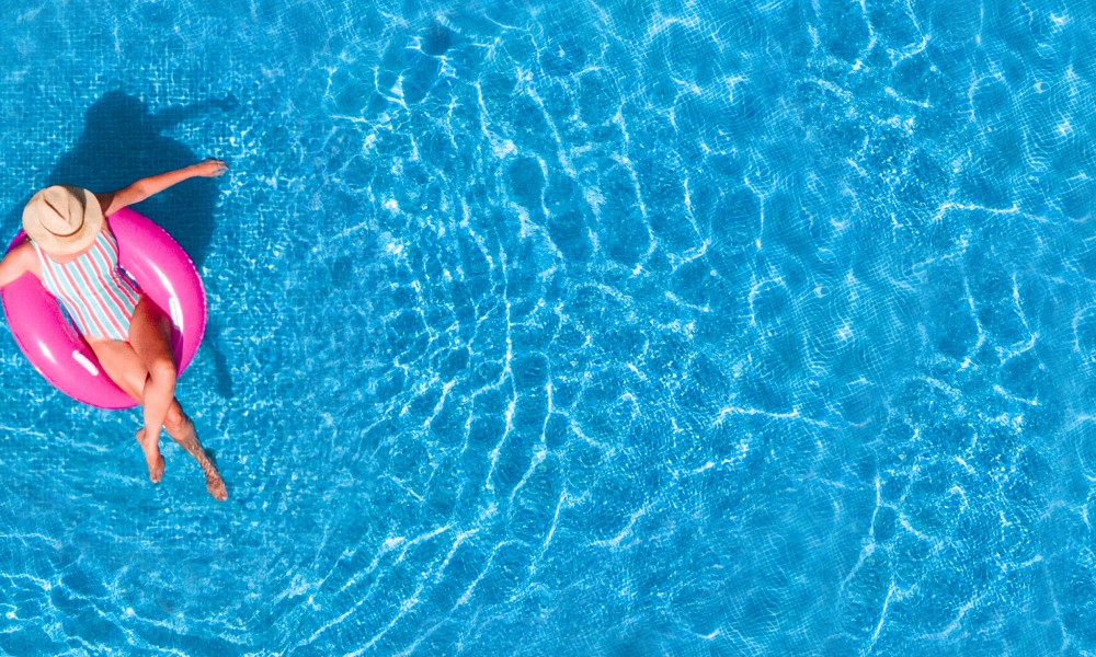 A woman sitting in a pink flotation ring in a pool wearing a multicolor swimsuit with vertical stripes and a sun hat.