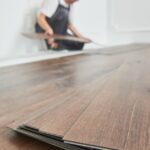 A stack of luxury vinyl flooring planks is in focus in the foreground, and a worker is installing the floor in the background.
