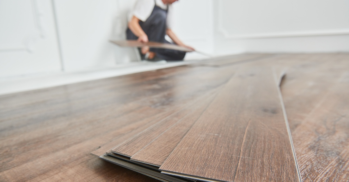 A stack of luxury vinyl flooring planks is in focus in the foreground, and a worker is installing the floor in the background.