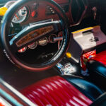 Interior view of a vehicle's front seat. The vehicle has red leather seats, a black steering wheel, and a cooler in passenger's seat.