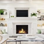 A luxurious home living room with a large fireplace in the center surrounded by white accents, decor, and furniture.