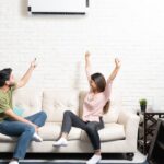 Mini Split vs. Central Air Conditioning: Which is Better? A man and woman sit on a couch. The man uses a remote to control a mini split above them while the woman cheers.