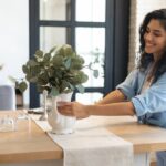 The Dos and Don’ts of Decorating Your Dining Room A smiling young woman setting a vase containing a broad-leafed plant on top of a wooden dining table.