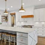 Farmhouse Style 101: The Core Design Elements A wide view of a modern farmhouse kitchen, including white cabinetry, a large white counter island, and open shelving.