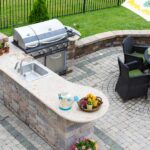 Renovation Tips: Getting Your Home Ready for Summer A patio in the backyard of a home with a grill, countertop, and table for dining in front of a black fence.