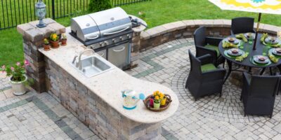 A patio in the backyard of a home with a grill, countertop, and table for dining in front of a black fence.